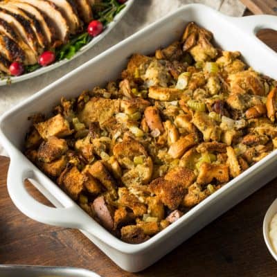 Golden gluten-free stuffing (classic or cornbread) in a baking dish showing a crispy top and fresh herbs surrounded by a platter of sliced turkey, mashed potatoes, gravy, and cranberry sauce