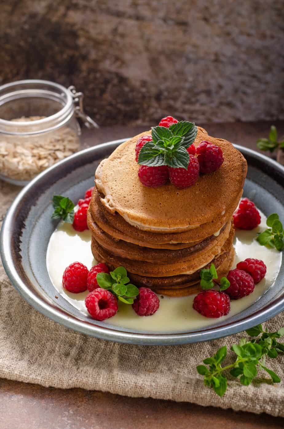 A high stack of pancakes with fresh raspberries and mint leaves on top and scattered around the pancakes on the plate and a jar of oat flour in the background