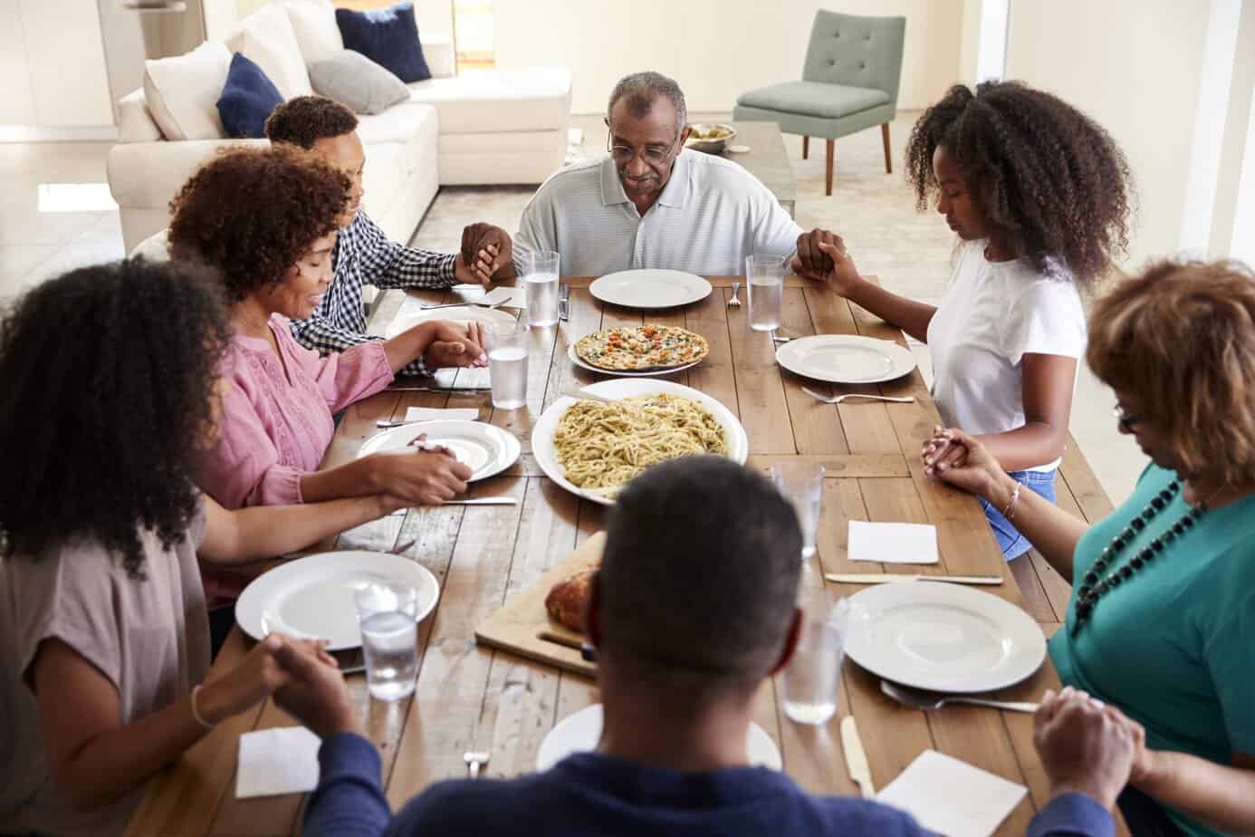 Three generation family holding hands and saying grace at wooden table before dinner, elevated view with a white sofa and green upholstered chair in the background