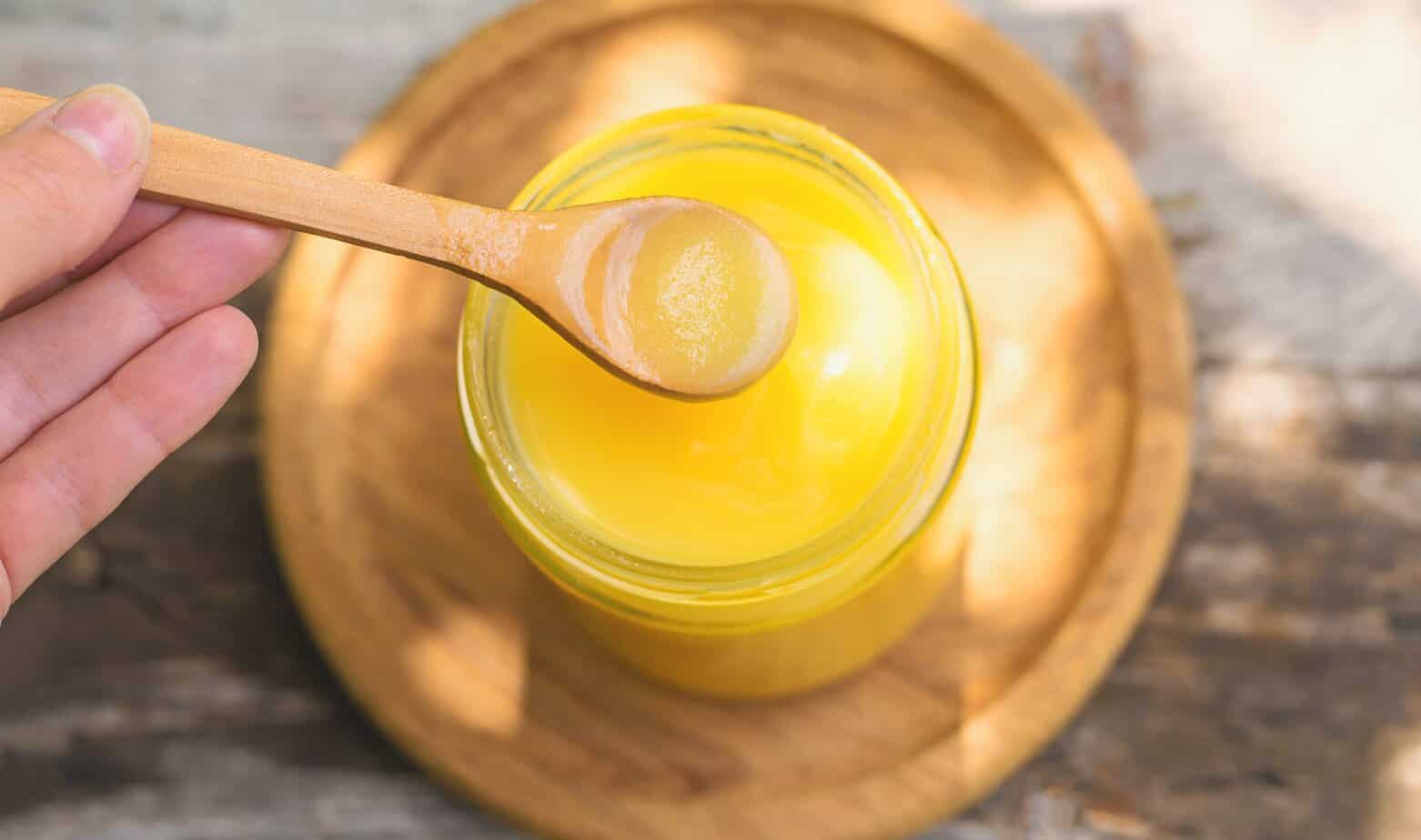 Ghee in a jar in a meadow. Selective focus. Ghee in a glass jar on a round wooden tray with a hand holding wooden spoon over the jar with some ghee in the spoon and a blurred background