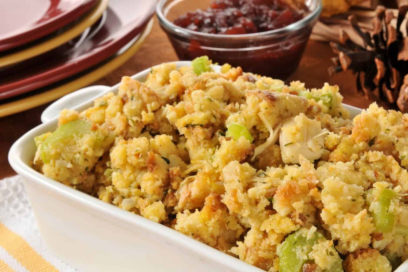 Landscape Images - Cornbread stuffing A casserole dish filled with gluten-free cornbread stuffing mixed with celery and onions, with cranberry sauce and stacked plates in the background