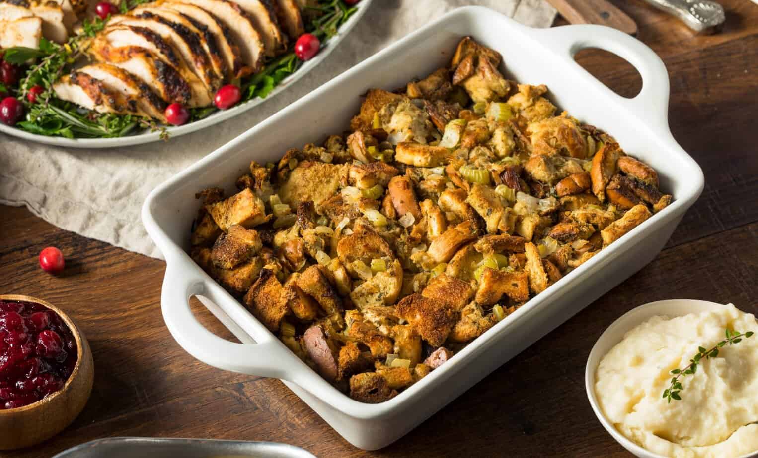 Homemade Thanksgiving Turkey Stuffing Golden gluten-free stuffing (classic or cornbread) in a baking dish showing a crispy top and fresh herbs surrounded by a platter of sliced turkey, mashed potatoes, gravy, and cranberry sauce
