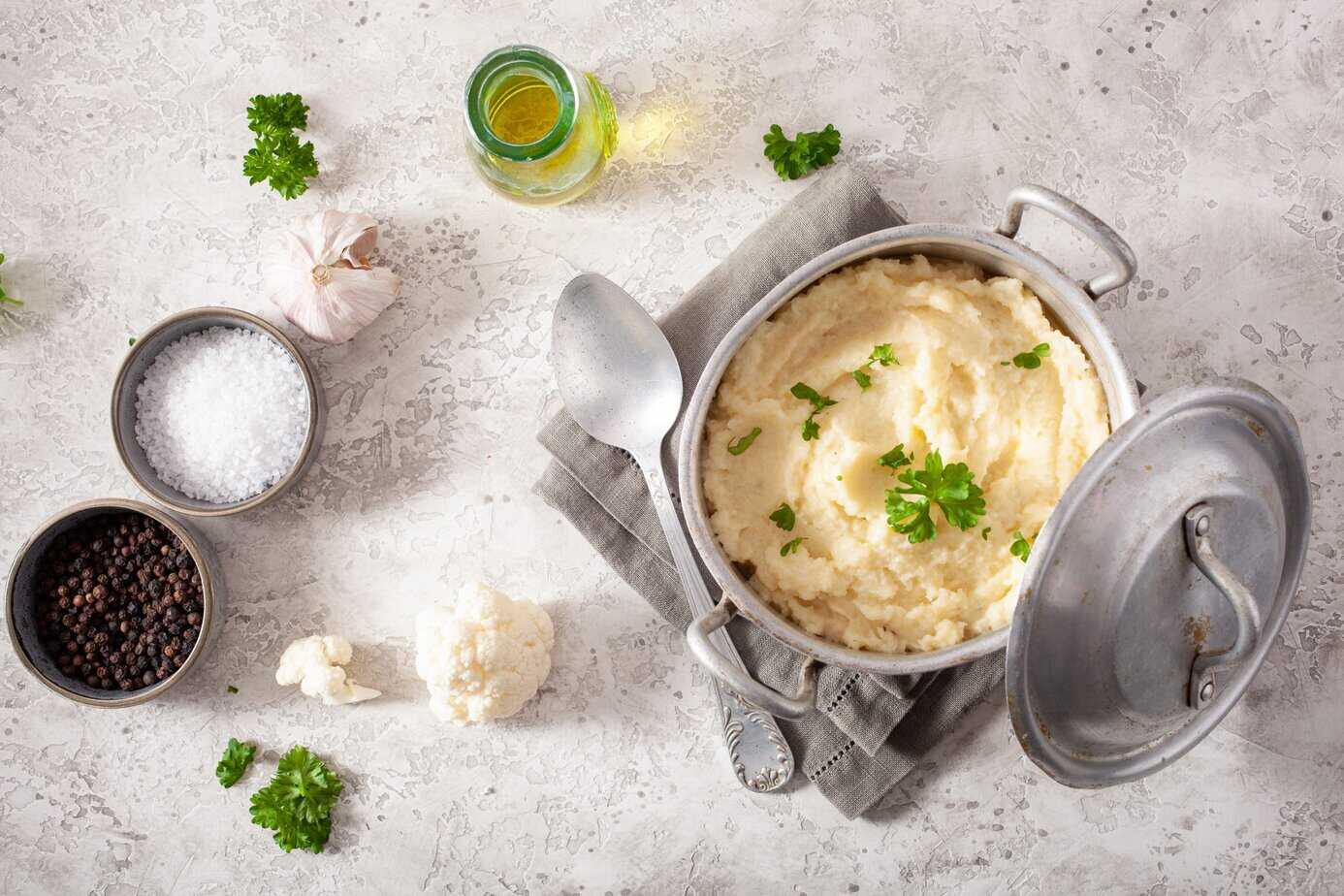 mashed cauliflower with butter. ketogenic paleo diet side dish A rustic metal pot filled with creamy mashed cauliflower garnished with fresh parsley, sitting on a gray linen napkin. Surrounding the pot are ingredients including coarse salt, black peppercorns, garlic, cauliflower florets, olive oil, and scattered parsley leaves on a textured light-gray surface