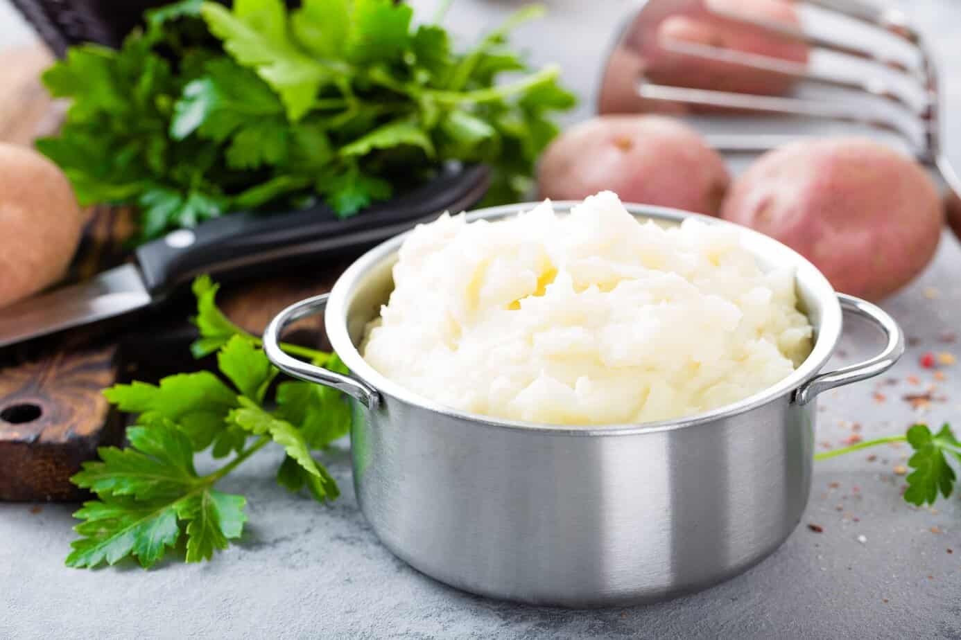 Mashed, mash potato with butter and milk. Potato puree A stainless steel pot filled with fluffy mashed potatoes sits on a light countertop, surrounded by fresh parsley, whole red potatoes, a knife, and a potato masher