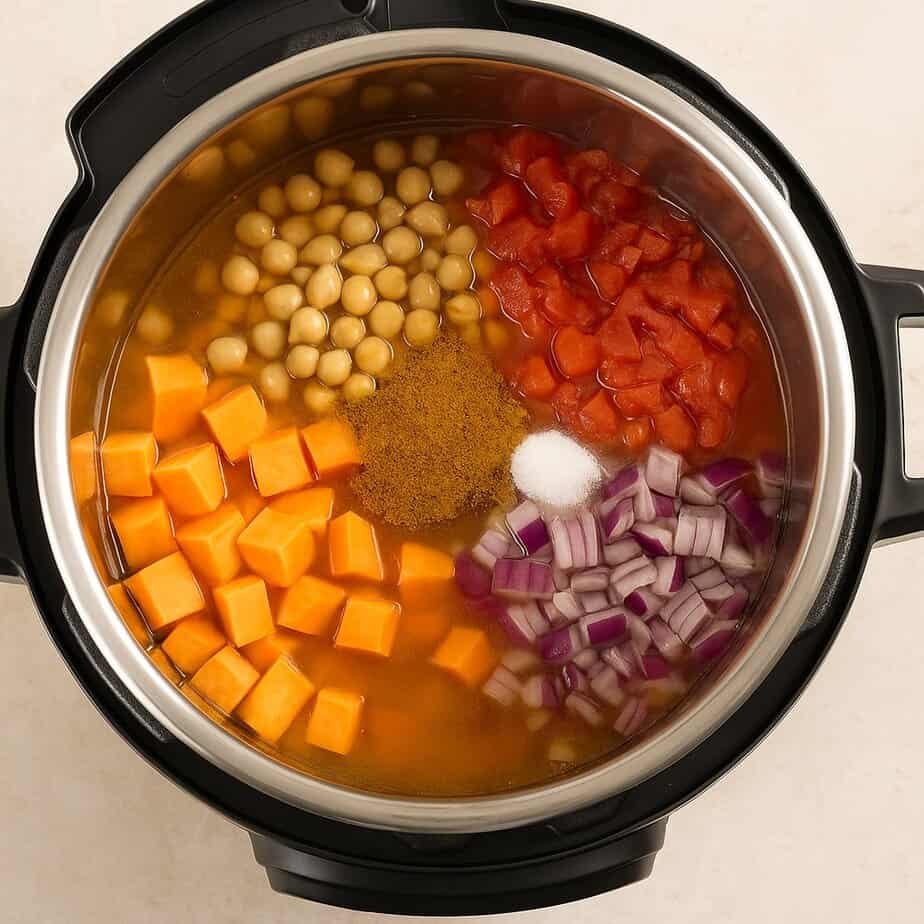 Overhead view of an Instant Pot filled with uncooked ingredients: cubed sweet potatoes, soaked chickpeas, chopped red onion, diced tomatoes, chicken broth, Ras El Hanout, salt, and baking soda, all arranged in colorful sections before cooking