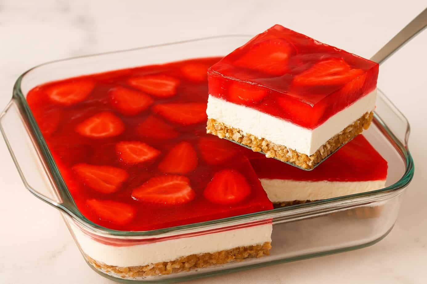 Strawberry Pretzel Salad A glass 8×8-inch baking dish filled with strawberry pretzel salad sits on a light countertop. One square piece is being lifted out with a metal spatula, showing the dessert’s three layers: a crisp pretzel crust on the bottom, a thick creamy middle layer, and a glossy strawberry gelatin topping with sliced strawberries suspended inside