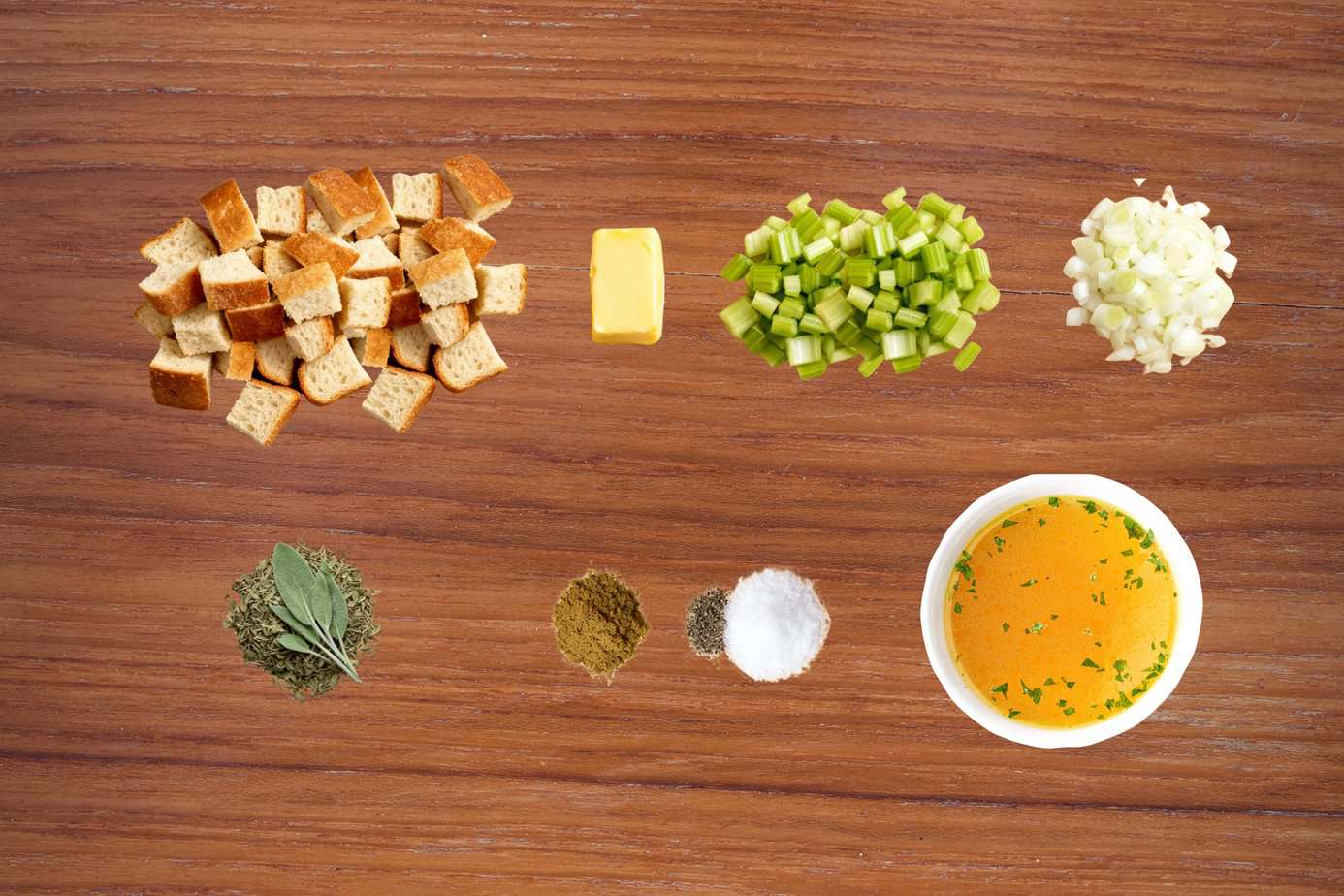 Landscape Images - Stuffing-ingredient-flat-lay A flat-lay image on a wooden surface showing the ingredients for classic turkey stuffing: dried gluten-free bread cubes, a pat of butter, chopped celery, chopped onions, dried and fresh sage, poultry seasoning, salt, pepper, and a bowl of chicken broth