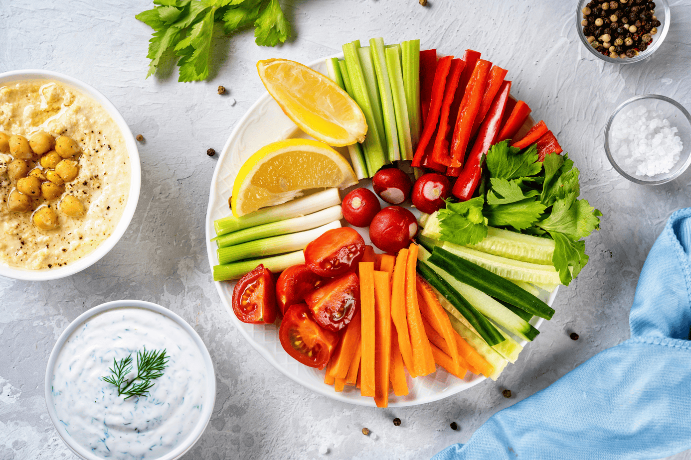 Veggie tray with hummus and Dilliscious A colorful vegetable platter arranged in a semi-circle with sliced cucumbers, carrots, red bell peppers, celery, radishes, cherry tomatoes, and lemon wedges, served alongside bowls of hummus and Dilliscious dip on a light gray surface