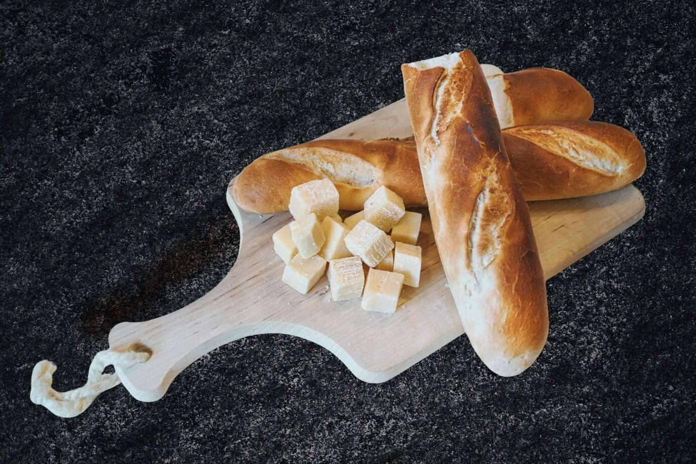 Landscape Images - cutting-baguette-bread-cubes A wooden cutting board on a dark countertop holding several crusty baguettes and a pile of evenly cut bread cubes, ready to be dried for homemade stuffing