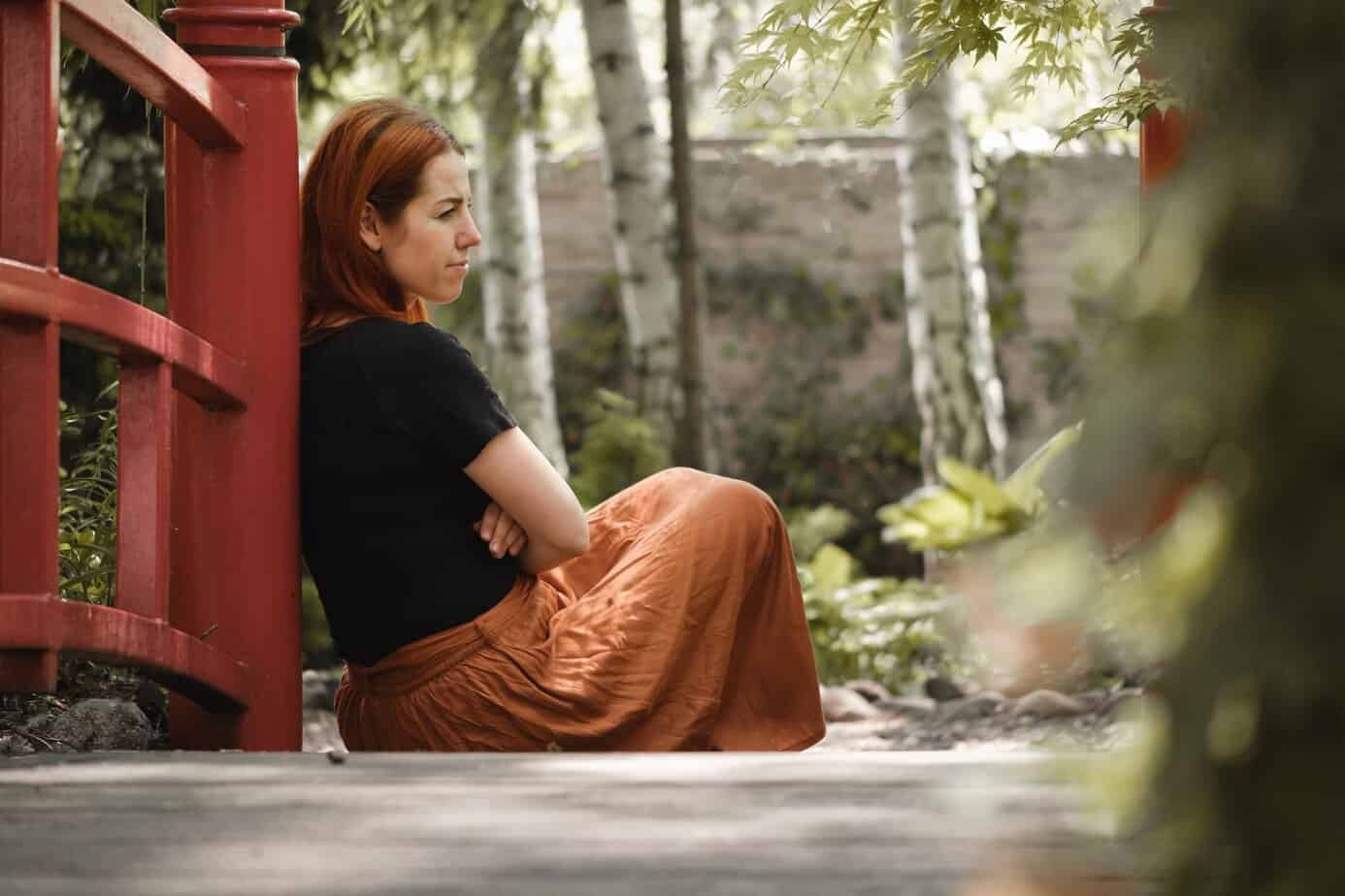 Woman sitting outdoors near a red bridge, seated with arms folded and looking away in a quite reflective posture