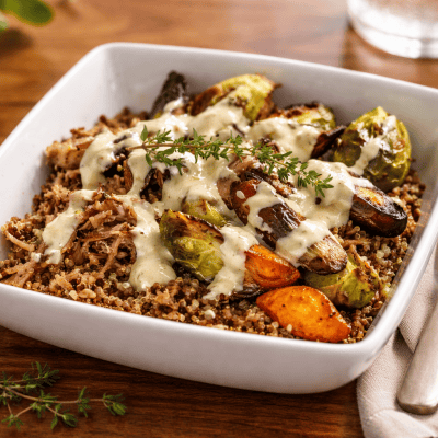 Brisket bowl with roasted Brussels sprouts and rainbow carrots over quinoa, drizzled with lemon tahini sauce and topped with fresh thyme, served in a white square bowl on a wooden table with a linen napkin and fork