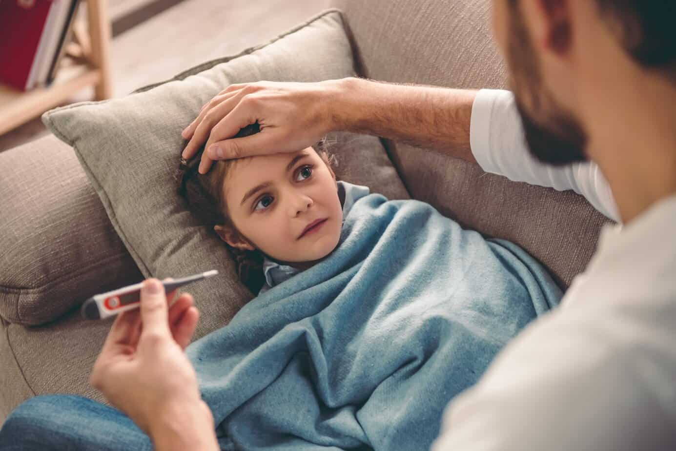 Little girl having a cold Parent checking a child’s temperature with a thermometer while the child rests under a blanket on a couch, illustrating caregiver concern during fever