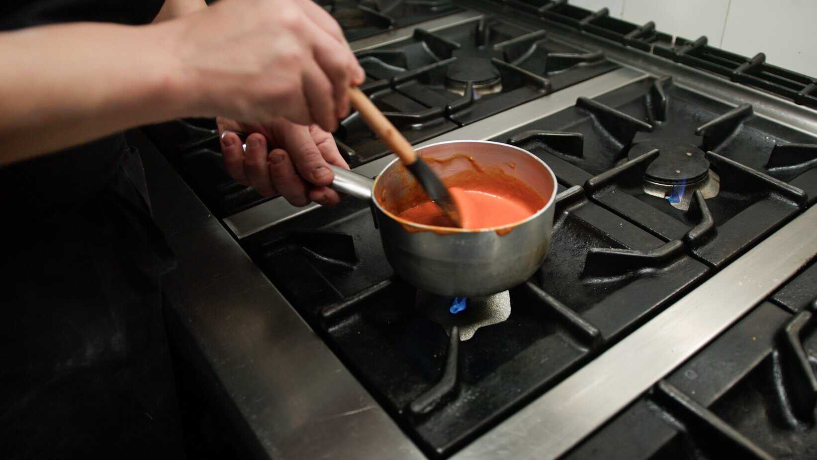 Person whisking bright orange buffalo sauce in a saucepan on a gas stove