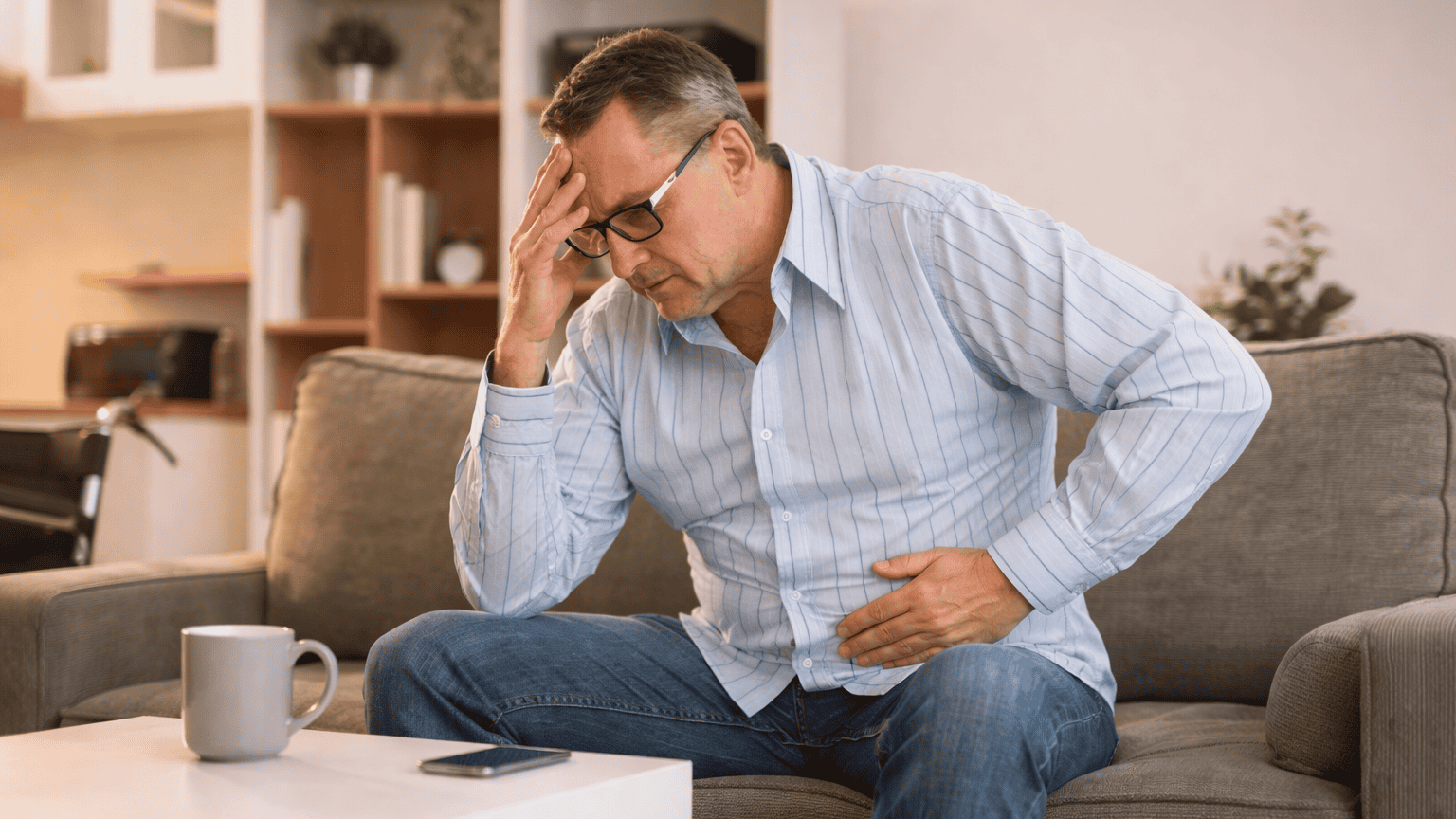 Middle-aged-man-with-chronic-inflammation Middle-aged man sitting on a couch holding his head and abdomen, illustrating fatigue and digestive discomfort associated with dysbiosis, leaky gut, and inflammation