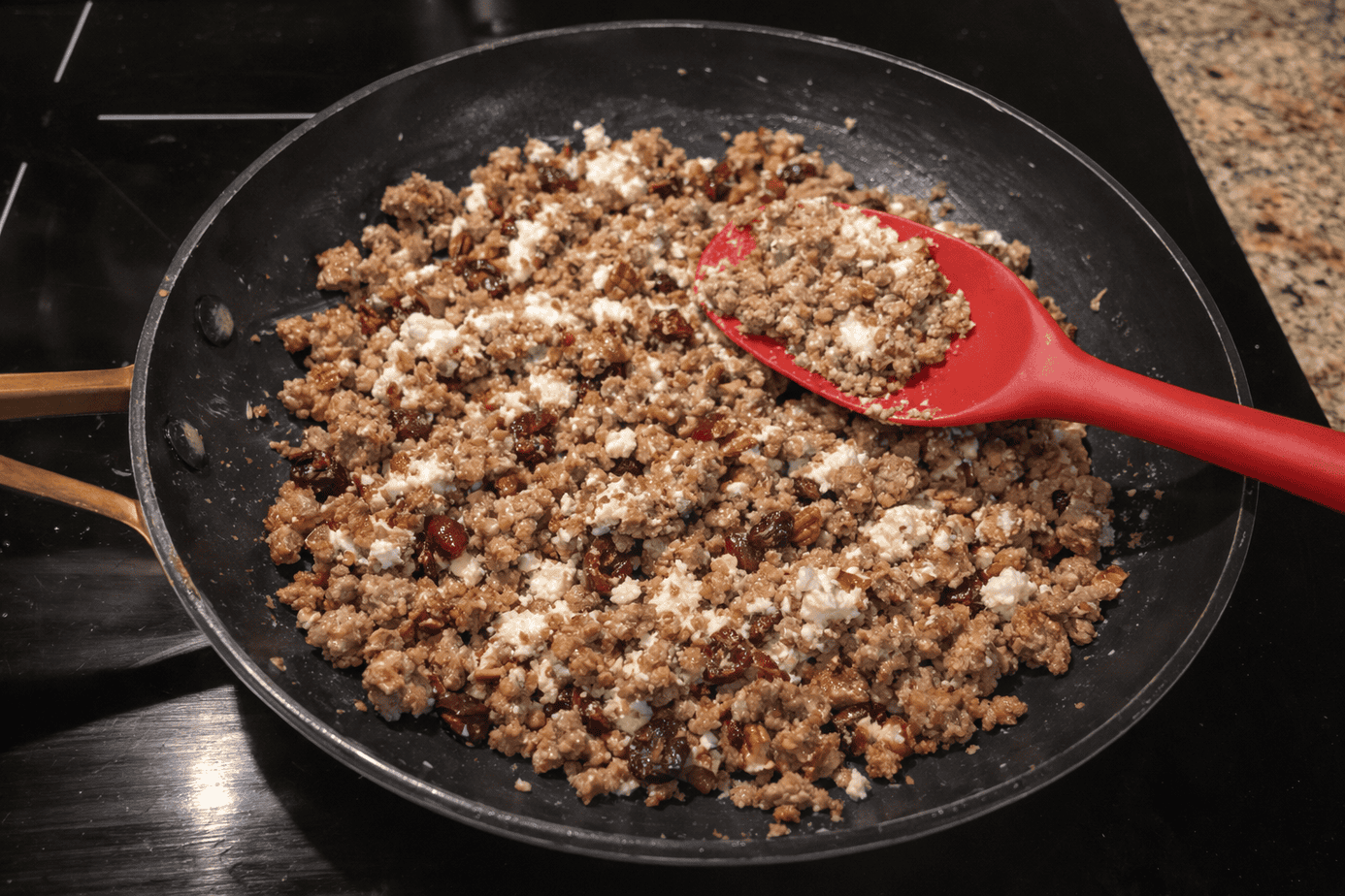 Ground pork sausage stuffing with pecans, cranberries, and goat cheese cooking in a skillet with a red silicon spoon