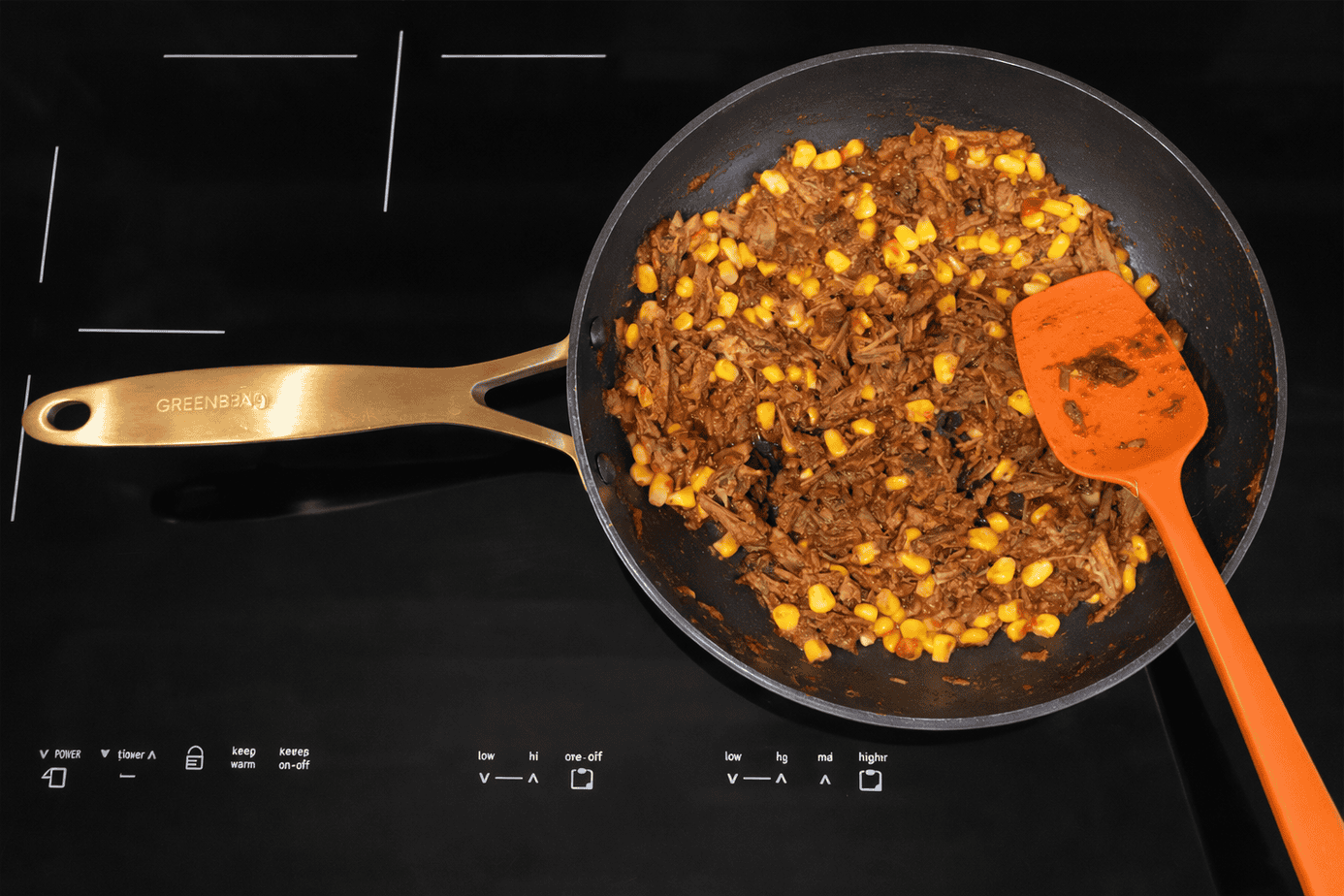 Top-down view of shredded beef and corn simmering in a black non-stick skillet on a glossy black induction cooktop, with an orange silicone spatula resting in the pan