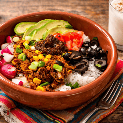 Shredded beef and rice taco bowl topped with avocado, tomatoes, radishes, black olives, green onions, and roasted salsa in a rustic terracotta bowl on a wooden table with a Mexican-style napkin and glass of horchata