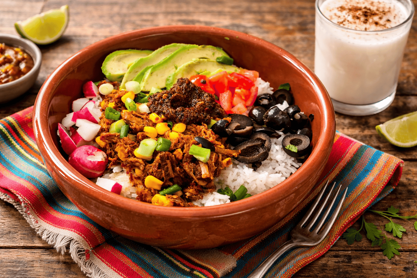Shredded beef and rice taco bowl topped with avocado, tomatoes, radishes, black olives, green onions, and roasted salsa in a rustic terracotta bowl on a wooden table with a Mexican-style napkin and glass of horchata