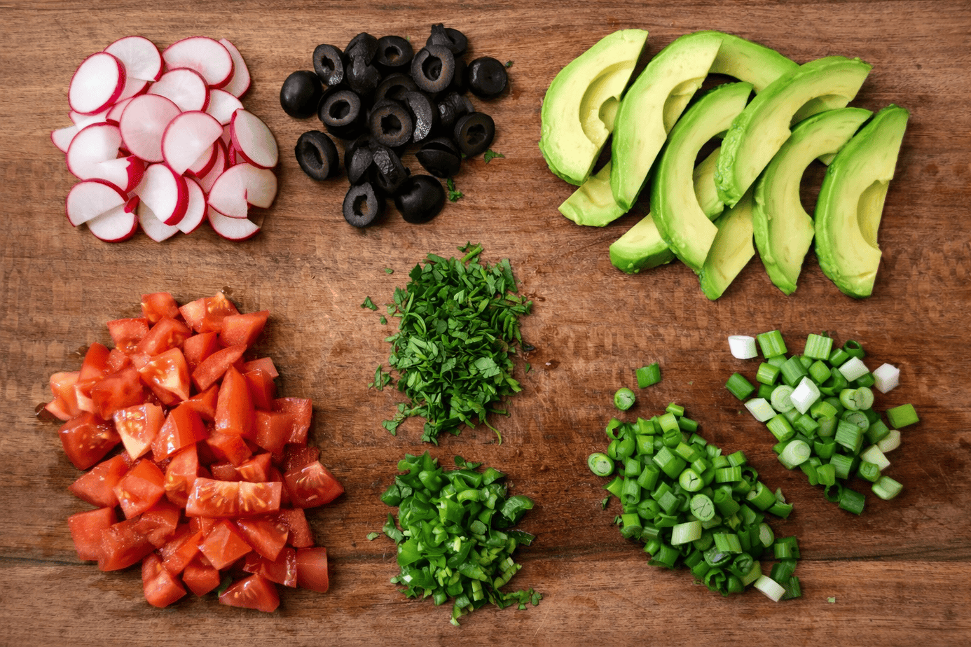 Prepped taco bowl ingredients arranged on a wooden cutting board, including sliced radishes, black olives, fresh avocado, diced tomatoes, chopped cilantro, jalapeños, and green onions