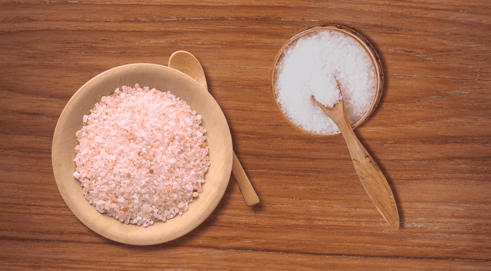 Himalayan pink and sea salt Two small wooden bowls filled with coarse salt, one with pink Himalayan salt and one with grayish-white sea salt, on a light wood surface with wooden spoons