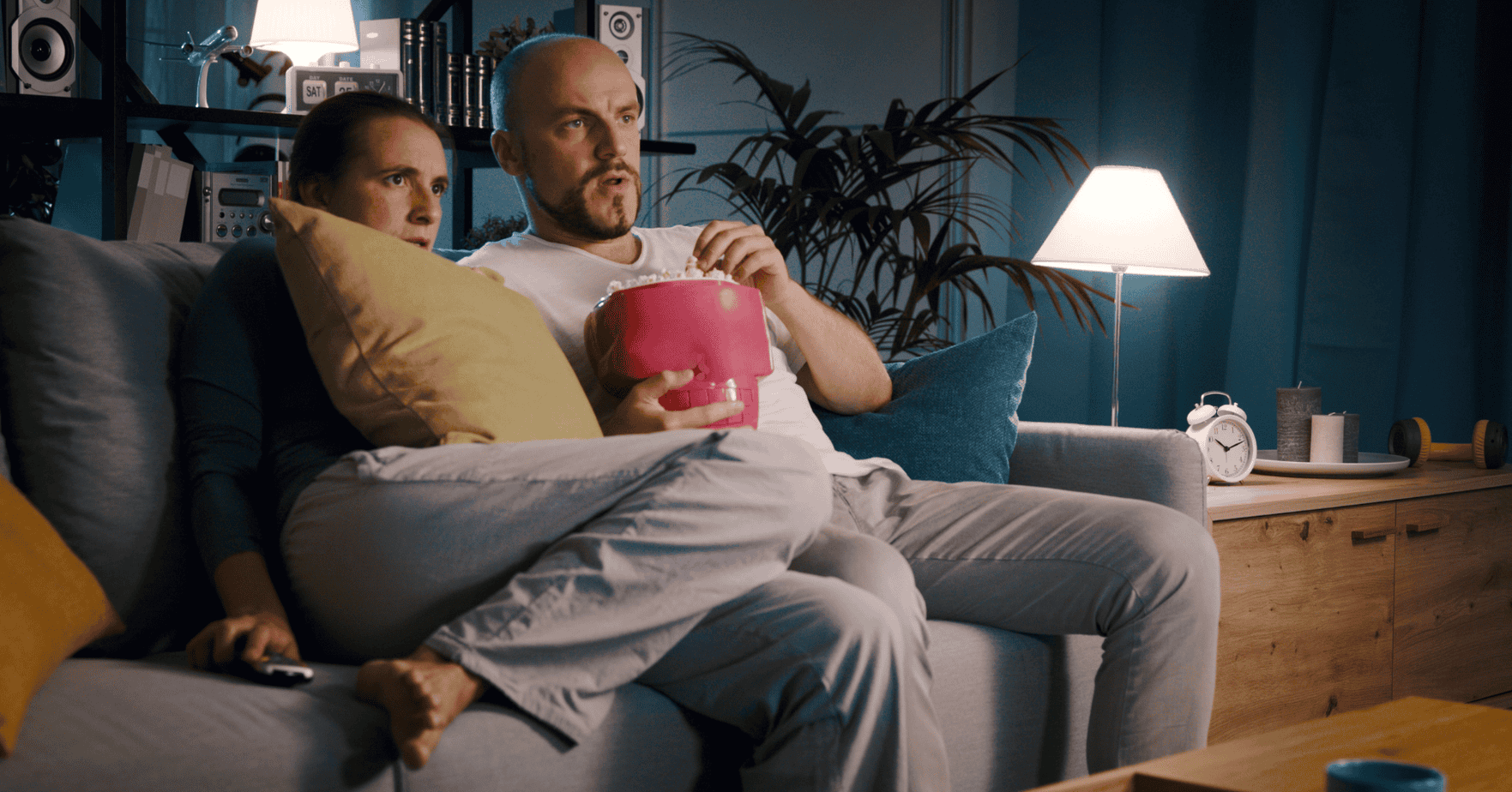 Couple watching a suspenseful movie together on the couch at night, woman clutching a pillow with a fearful expression while man reaches into a popcorn bowl