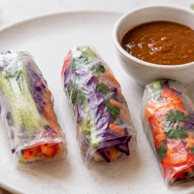 Three rainbow salad rolls with colorful vegetable fillings showing through rice paper wrappers on a white plate with almond butter dipping sauce
