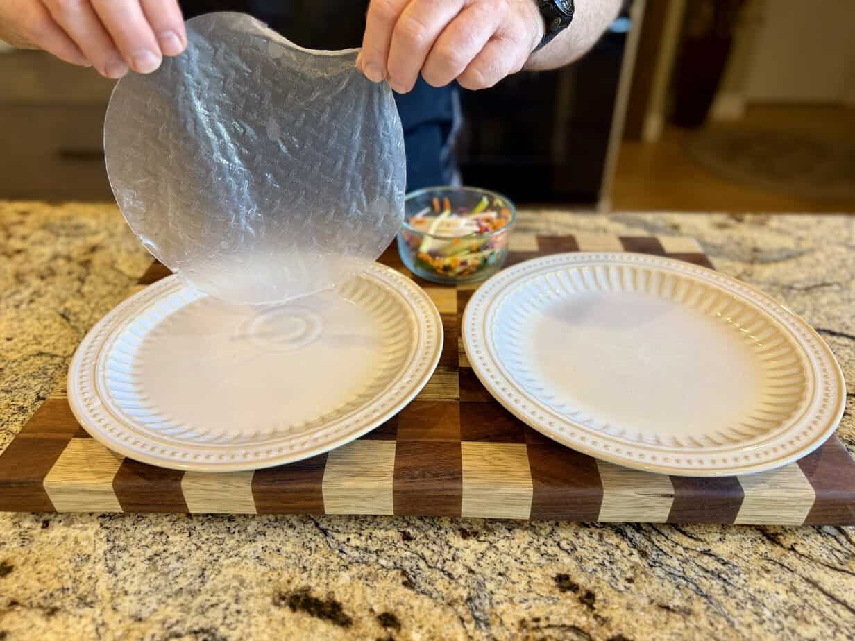 Dripping rice paper wrapper after dipping Hands holding a softened rice paper wrapper above a plate to allow excess water to drip off before assembling fresh rainbow salad rolls.