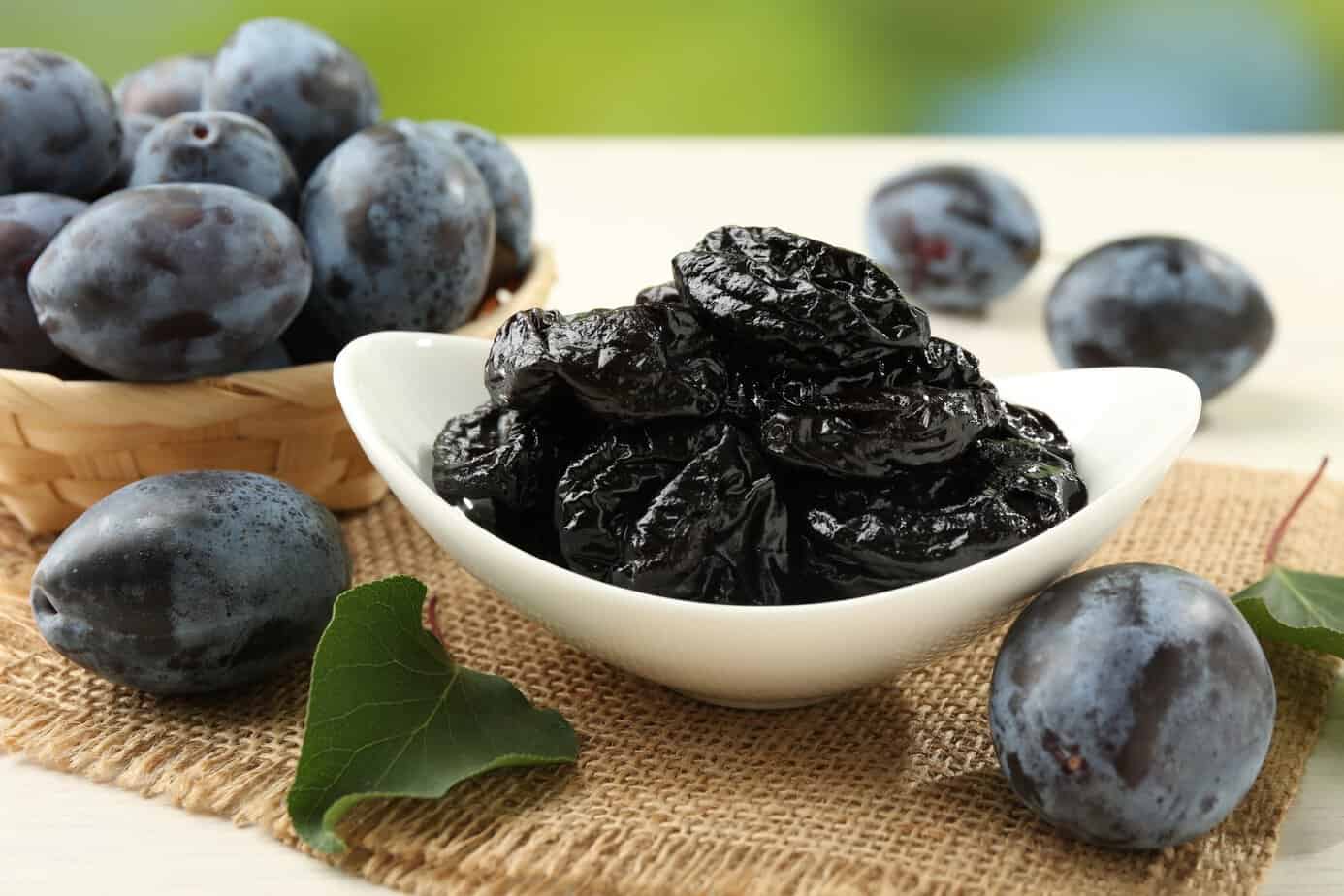 Dried prunes and fresh plums on table, closeup Bowl of dried prunes surrounded by fresh purple plums on a tan placemat and table